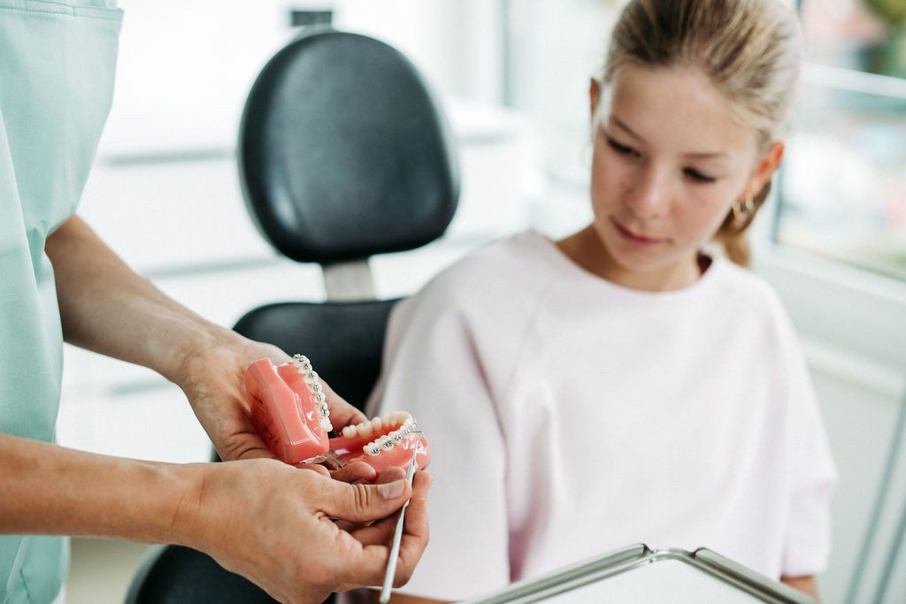 Eine Kieferorthopädin in mintfarbener Praxiskleidung hält ein Zahnmodell mit festsitzender Zahnspange in den Händen und erklärt es einer jungen Patientin. Die Patientin, ein Mädchen im Teenageralter mit blondem Haar, trägt ein hellrosa Oberteil und schaut interessiert auf das Modell. Im Hintergrund ist ein moderner, heller Behandlungsraum zu sehen. Die Szene vermittelt Beratung, Aufklärung und Vertrauen in einer freundlichen Praxisatmosphäre.