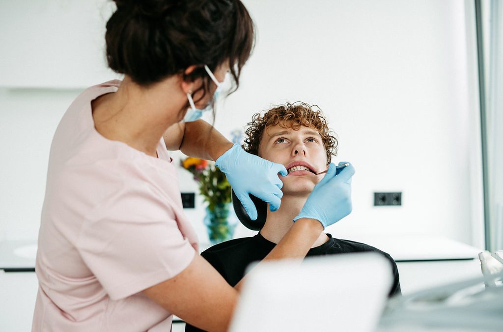 Eine Kieferorthopädin in rosa Praxiskleidung und mit blauen Handschuhen führt bei einem jugendlichen Patienten eine Untersuchung oder Behandlung durch. Der Jugendliche sitzt im Behandlungsstuhl mit leicht geöffnetem Mund, während die Behandlerin mit einem Instrument die Zähne inspiziert – möglicherweise im Rahmen einer Bracketumfeldversiegelung. Die Szene spielt in einem hellen, modernen Behandlungsraum. Im Hintergrund steht ein bunter Blumenstrauß, der eine freundliche Atmosphäre schafft.