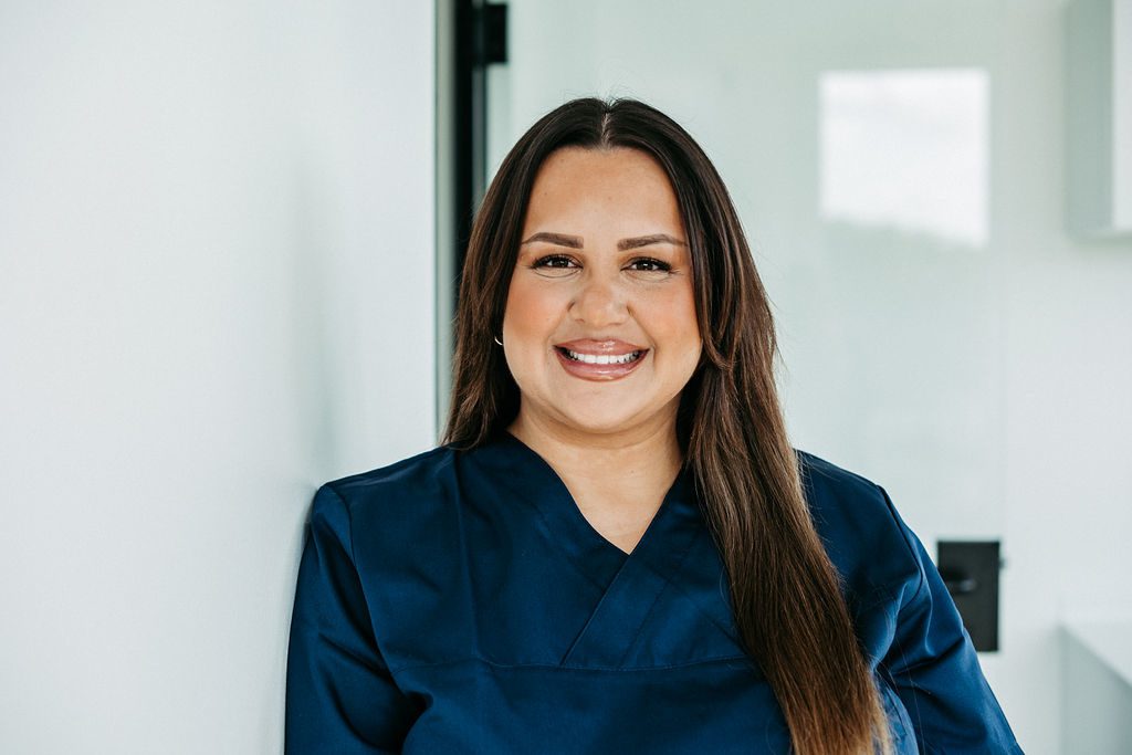 Eine Frau mit langen, glatten braunen Haaren steht in einer modernen, hellen Praxisumgebung und lächelt direkt in die Kamera. Sie trägt dunkelblaue Praxiskleidung und wirkt freundlich und professionell. Ihre offene Ausstrahlung vermittelt Herzlichkeit – passend zu ihrer Tätigkeit am Empfang und in der Assistenz.