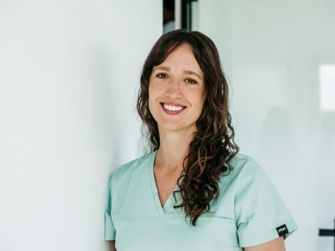 Eine Frau mit langen, lockigen braunen Haaren steht lächelnd an eine weiße Wand gelehnt in einer modernen, hellen Praxisumgebung. Sie trägt mintfarbene Praxiskleidung und blickt freundlich in die Kamera. Ihr offenes Lächeln und die entspannte Körperhaltung vermitteln Kompetenz und Herzlichkeit – passend zu ihrer Rolle als Kieferorthopädin.