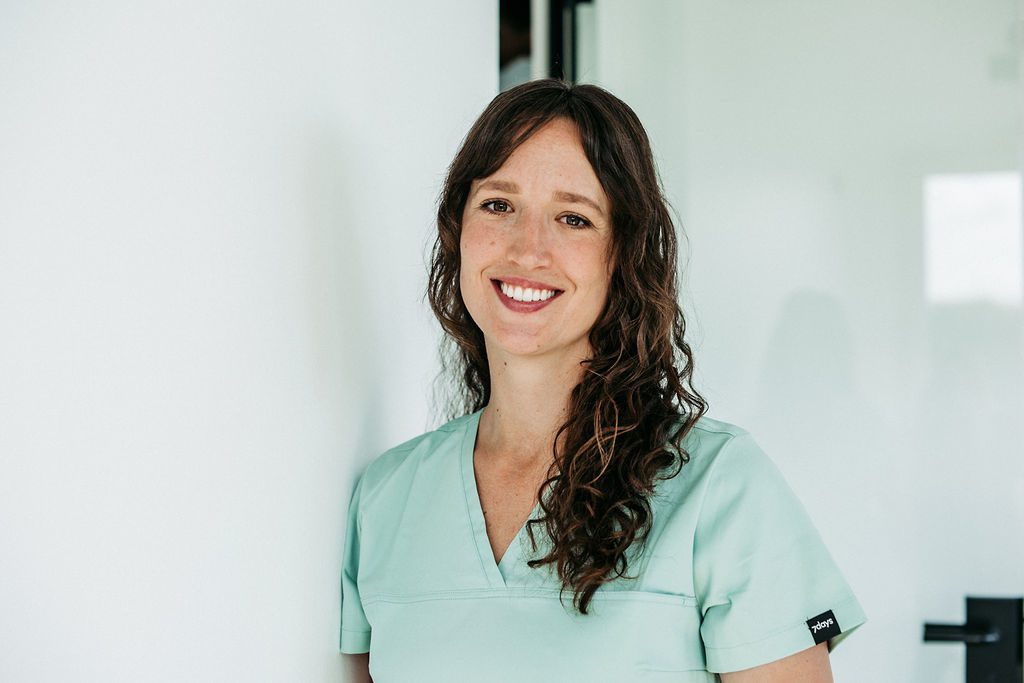 Eine Frau mit langen, lockigen braunen Haaren steht lächelnd an eine weiße Wand gelehnt in einer modernen, hellen Praxisumgebung. Sie trägt mintfarbene Praxiskleidung und blickt freundlich in die Kamera. Ihr offenes Lächeln und die entspannte Körperhaltung vermitteln Kompetenz und Herzlichkeit – passend zu ihrer Rolle als Kieferorthopädin.