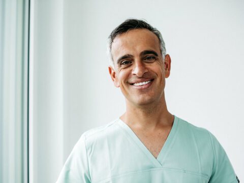 Ein Mann mittleren Alters mit kurzen, dunkelgrau melierten Haaren steht in einer hellen, modernen Praxisumgebung nahe eines Fensters. Er trägt mintfarbene Praxiskleidung und lächelt freundlich in die Kamera. Seine entspannte und offene Ausstrahlung vermittelt Kompetenz und Vertrauen – passend zu seiner Rolle als Kieferorthopäde.