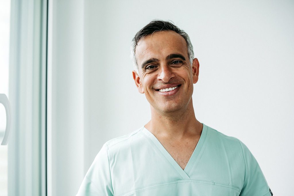 Ein Mann mittleren Alters mit kurzen, dunkelgrau melierten Haaren steht in einer hellen, modernen Praxisumgebung nahe eines Fensters. Er trägt mintfarbene Praxiskleidung und lächelt freundlich in die Kamera. Seine entspannte und offene Ausstrahlung vermittelt Kompetenz und Vertrauen – passend zu seiner Rolle als Kieferorthopäde.