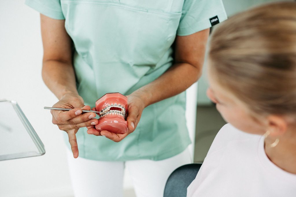 Eine Person in mintfarbener Praxiskleidung erklärt einer jugendlichen Patientin mithilfe eines kieferorthopädischen Modells mit festsitzender Zahnspange den Ablauf oder die Wirkung der Behandlung. Mit einem Instrument wird auf bestimmte Stellen am Modell gezeigt. Die Patientin sitzt seitlich im Behandlungsstuhl und hört aufmerksam zu. Die Szene vermittelt Aufklärung, Vertrauen und professionelle Betreuung in der Kieferorthopädie.
