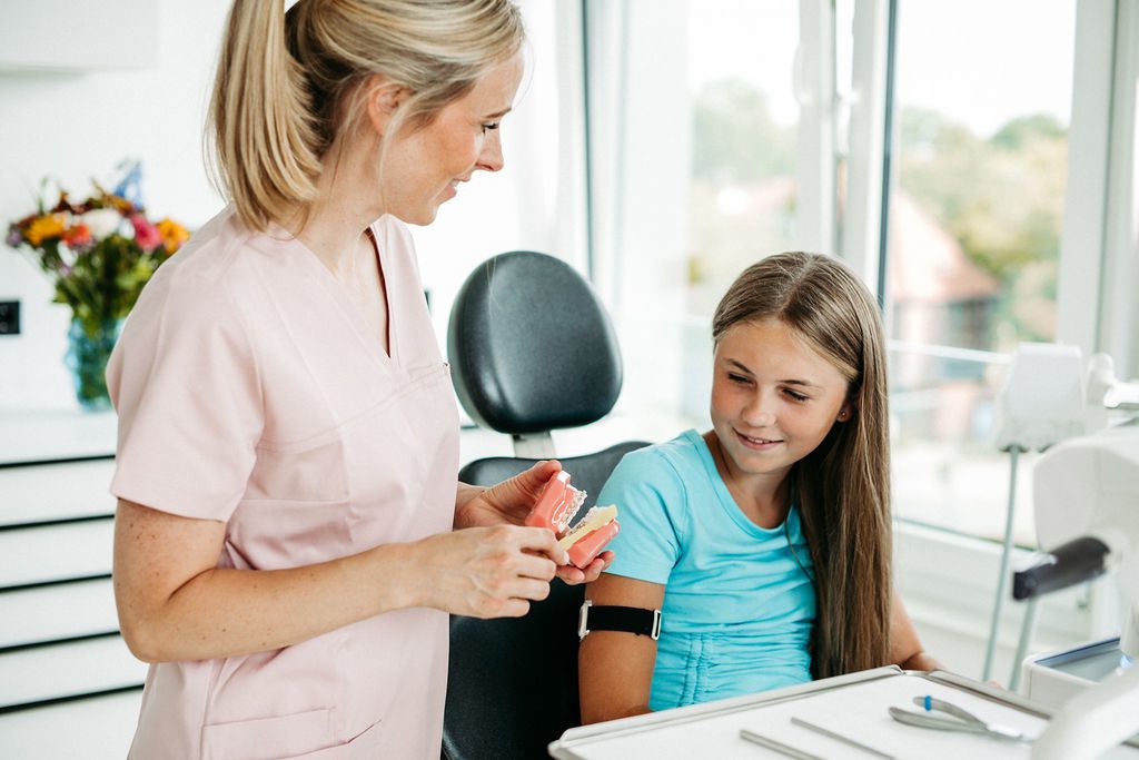 Eine Kieferorthopädin in rosa Praxiskleidung erklärt einem jungen Mädchen anhand eines Zahnmodells mit Zahnspange die Funktionsweise einer festen kieferorthopädischen Apparatur. Die Patientin sitzt im Behandlungsstuhl, hört aufmerksam zu und lächelt. Das Modell zeigt deutlich selbstligierende Brackets, die kleiner und ästhetischer wirken als herkömmliche Varianten. Im Hintergrund sind helle Fensterfronten und ein bunter Blumenstrauß zu sehen, was die freundliche Atmosphäre der Praxis unterstreicht.