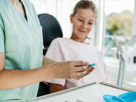 Eine Kieferorthopädin in mintfarbener Praxiskleidung hält eine herausnehmbare lose Zahnspange in den Händen und erklärt diese einem jungen Mädchen im Behandlungsstuhl. Das Mädchen trägt ein hellrosa Oberteil und schaut neugierig und lächelnd auf die Spange. Der helle Praxisraum im Hintergrund sorgt für eine freundliche, moderne Atmosphäre. Die Szene verdeutlicht die kindgerechte Beratung zu herausnehmbaren Zahnspangen.