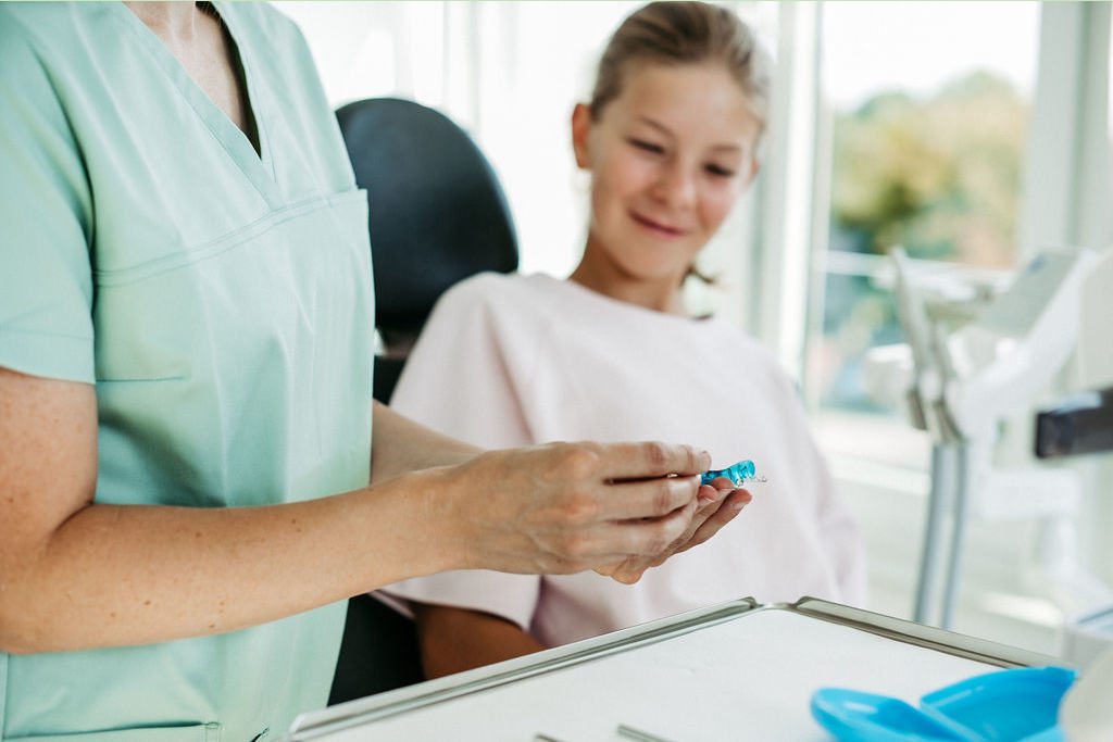 Eine Kieferorthopädin in mintfarbener Praxiskleidung hält eine herausnehmbare lose Zahnspange in den Händen und erklärt diese einem jungen Mädchen im Behandlungsstuhl. Das Mädchen trägt ein hellrosa Oberteil und schaut neugierig und lächelnd auf die Spange. Der helle Praxisraum im Hintergrund sorgt für eine freundliche, moderne Atmosphäre. Die Szene verdeutlicht die kindgerechte Beratung zu herausnehmbaren Zahnspangen.