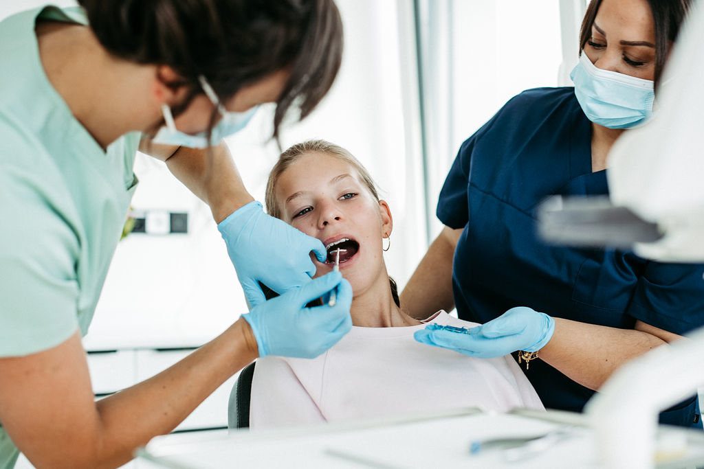 Eine Jugendliche sitzt auf einem Behandlungsstuhl in einer kieferorthopädischen Praxis. Eine Zahnärztin mit Mundschutz und blauen Handschuhen untersucht den geöffneten Mund des Mädchens mit einem Spiegelinstrument, während eine Assistentin in blauer Praxiskleidung unterstützend danebensteht. Das Mädchen trägt ein helles Oberteil. Die Atmosphäre ist hell, modern und professionell.