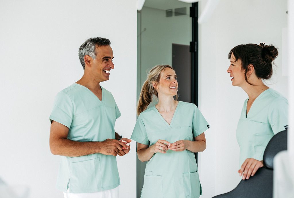Zwei Kieferorthopädinnen und ein Kieferorthopäde stehen gemeinsam in einem hellen Behandlungsraum. Alle tragen mintfarbene Praxiskleidung und unterhalten sich gut gelaunt. Die Szene strahlt Teamgeist, Kompetenz und eine freundliche, vertrauensvolle Atmosphäre aus – passend für eine moderne kieferorthopädische Praxis.
