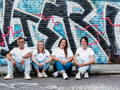 Vier Mitglieder des U-Smile-Teams sitzen entspannt nebeneinander auf dem Gehweg vor einer bunt besprühten Backsteinwand mit Graffiti. Alle tragen weiße T-Shirts und Jeans, was ein modernes, sympathisches und nahbares Teamgefühl vermittelt. Sie lächeln in die Kamera und strahlen Freundlichkeit sowie Teamgeist aus – passend für eine kieferorthopädische Praxis mit familiärer Atmosphäre.