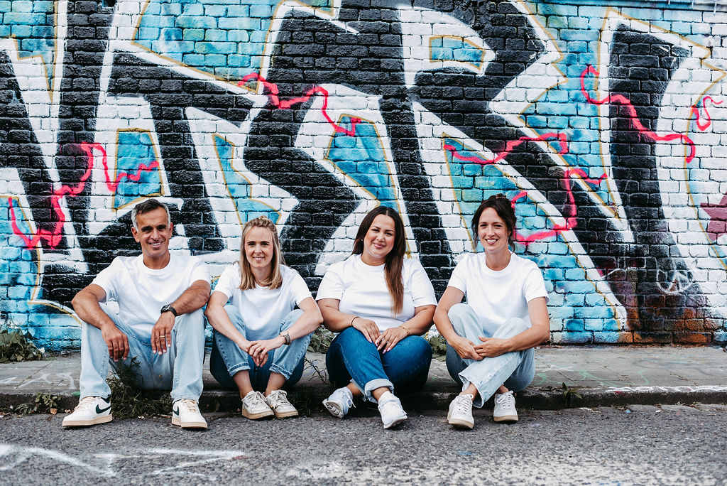 Vier Mitglieder des U-Smile-Teams sitzen entspannt nebeneinander auf dem Gehweg vor einer bunt besprühten Backsteinwand mit Graffiti. Alle tragen weiße T-Shirts und Jeans, was ein modernes, sympathisches und nahbares Teamgefühl vermittelt. Sie lächeln in die Kamera und strahlen Freundlichkeit sowie Teamgeist aus – passend für eine kieferorthopädische Praxis mit familiärer Atmosphäre.