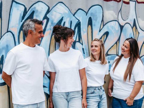 Vier Mitglieder des U-Smile Praxisteams stehen lachend nebeneinander vor einer Wand mit bunter Graffiti-Kunst. Alle tragen weiße T-Shirts und Jeans, was ein modernes und lockeres Teamgefühl vermittelt. Die Gruppe wirkt harmonisch, freundlich und offen – ein klares Zeichen für ein herzliches Miteinander und eine patientennahe Betreuung in der kieferorthopädischen Praxis.
