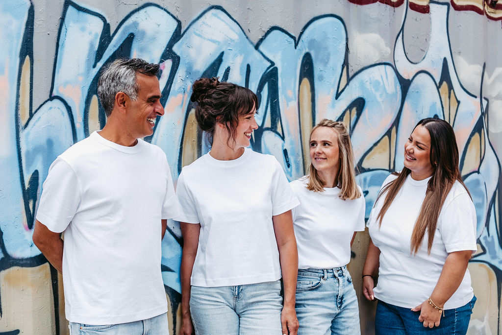Vier Mitglieder des U-Smile Praxisteams stehen lachend nebeneinander vor einer Wand mit bunter Graffiti-Kunst. Alle tragen weiße T-Shirts und Jeans, was ein modernes und lockeres Teamgefühl vermittelt. Die Gruppe wirkt harmonisch, freundlich und offen – ein klares Zeichen für ein herzliches Miteinander und eine patientennahe Betreuung in der kieferorthopädischen Praxis.