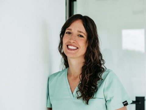 Eine Frau mit langen, braunen, lockigen Haaren steht lächelnd an eine weiße Wand gelehnt in einer modernen Praxisumgebung. Sie trägt mintfarbene Praxiskleidung und blickt freundlich in die Kamera. Der Hintergrund ist hell, mit Glaswänden und dezenten Spiegelungen, was eine offene, professionelle und einladende Atmosphäre vermittelt. Ihr sympathisches Auftreten lässt auf ein kompetentes und herzliches Praxisteam schließen.