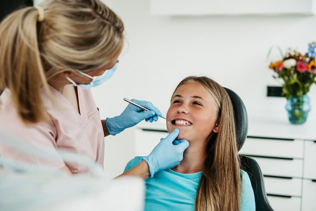 Ein junges Mädchen sitzt lächelnd auf einem Behandlungsstuhl in einer modernen kieferorthopädischen Praxis. Eine Kieferorthopädin in rosa Praxiskleidung, mit Schutzmaske und blauen Handschuhen, untersucht die Zähne des Mädchens vorsichtig mit einem Spiegelinstrument. Beide wirken entspannt, die Atmosphäre ist freundlich und vertrauensvoll. Im Hintergrund sorgt ein bunter Blumenstrauß in einer Vase für eine persönliche Note im hellen Praxisraum.