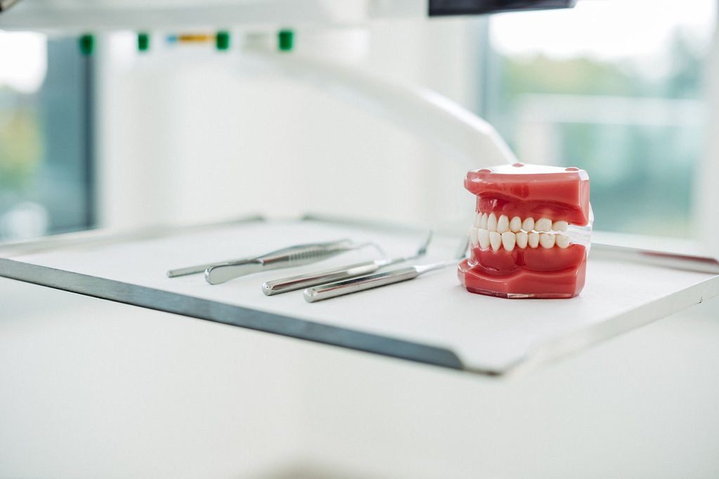 Auf einem Behandlungsablage-Tisch in einer modernen kieferorthopädischen Praxis liegen drei silberne zahnärztliche Instrumente ordentlich nebeneinander – darunter ein Mundspiegel, eine Pinzette und eine Sonde. Daneben steht ein Zahnmodell mit vollständigem Gebiss, das zur Veranschaulichung von Zahnstellungen und kieferorthopädischen Behandlungen dient. Im Hintergrund ist durch das Fenster unscharf die Umgebung außerhalb der Praxis zu erkennen. Die Szene wirkt steril, professionell und gut vorbereitet für die Behandlung.