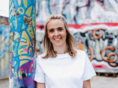 Eine junge Frau mit schulterlangem blondem Haar steht draußen vor einer bunt besprühten Wand mit urbanem Graffiti. Sie trägt ein weißes T-Shirt und lehnt sich locker an eine farbenfroh bemalte Säule. Die Frau lächelt selbstbewusst in die Kamera – ihre geraden Zähne deuten auf eine abgeschlossene kieferorthopädische Behandlung hin. Die Szene vermittelt Lebensfreude, Selbstsicherheit und Stabilität – passend zum Thema Retainer.