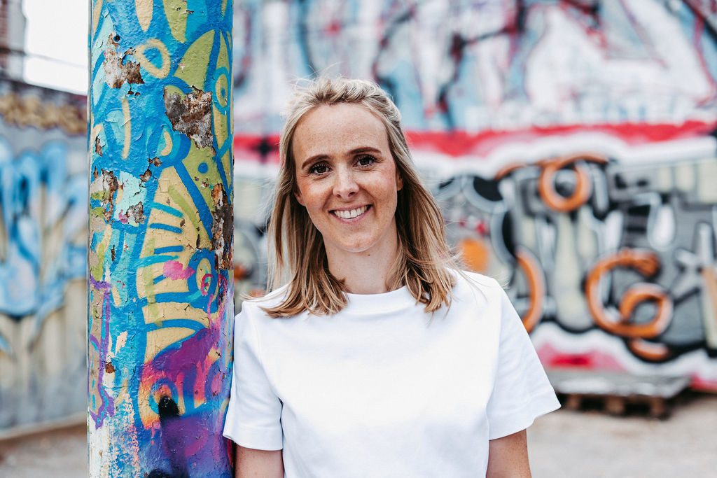 Eine junge Frau mit schulterlangem blondem Haar steht draußen vor einer bunt besprühten Wand mit urbanem Graffiti. Sie trägt ein weißes T-Shirt und lehnt sich locker an eine farbenfroh bemalte Säule. Die Frau lächelt selbstbewusst in die Kamera – ihre geraden Zähne deuten auf eine abgeschlossene kieferorthopädische Behandlung hin. Die Szene vermittelt Lebensfreude, Selbstsicherheit und Stabilität – passend zum Thema Retainer.