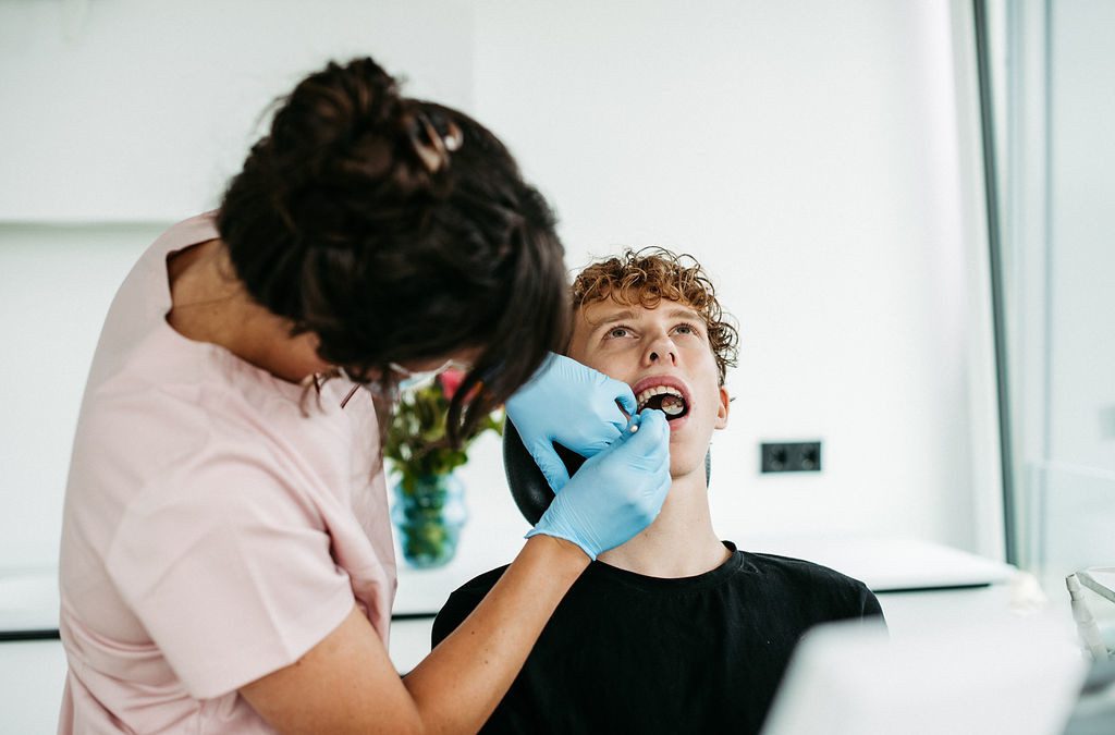 Ein Jugendlicher sitzt mit geöffnetem Mund auf einem Behandlungsstuhl in einer modernen kieferorthopädischen Praxis. Eine Kieferorthopädin in rosa Praxiskleidung und blauen Handschuhen untersucht sorgfältig die Innenseite seines Oberkiefers – ein Hinweis auf die Anwendung der Lingualtechnik, bei der die Zahnspange unsichtbar auf der Innenseite der Zähne befestigt wird. Der Raum wirkt hell und freundlich, im Hintergrund steht ein bunter Blumenstrauß.