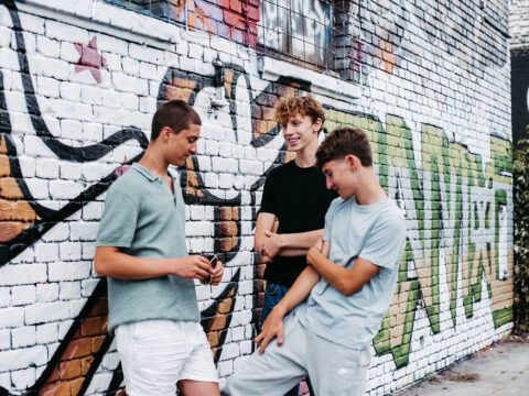 Drei jugendliche Jungen stehen locker vor einer farbenfroh besprühten Backsteinwand mit Graffiti. Sie lachen und unterhalten sich entspannt, einer hält eine Sonnenbrille in der Hand. Alle drei tragen lässige Freizeitkleidung. Ihre Zähne wirken gepflegt und gerade – ein möglicher Hinweis auf die Anwendung von unsichtbaren Alignern. Die Szene vermittelt Selbstbewusstsein, Gemeinschaft und jugendliche Leichtigkeit im urbanen Umfeld.