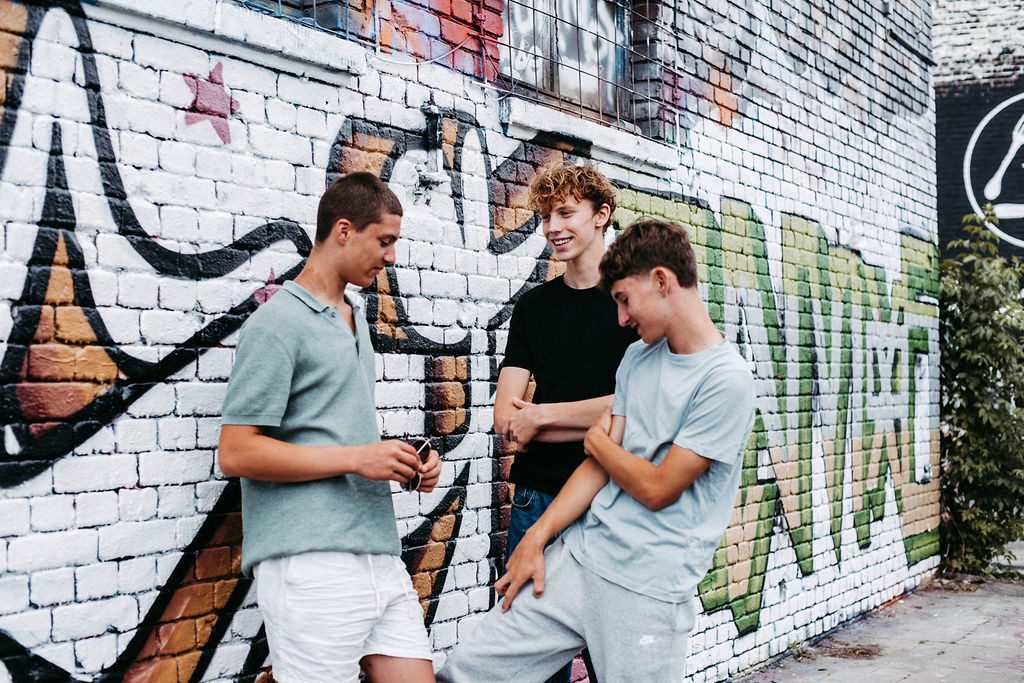 Drei jugendliche Jungen stehen locker vor einer farbenfroh besprühten Backsteinwand mit Graffiti. Sie lachen und unterhalten sich entspannt, einer hält eine Sonnenbrille in der Hand. Alle drei tragen lässige Freizeitkleidung. Ihre Zähne wirken gepflegt und gerade – ein möglicher Hinweis auf die Anwendung von unsichtbaren Alignern. Die Szene vermittelt Selbstbewusstsein, Gemeinschaft und jugendliche Leichtigkeit im urbanen Umfeld.