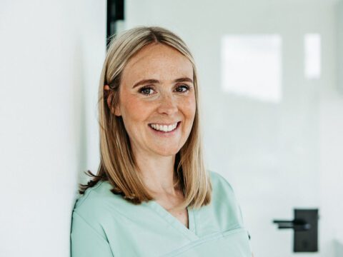 Dr. Alexandra Lampen, eine Frau mit schulterlangem, glattem, blondem Haar, steht in einem hellen, modern eingerichteten Praxisraum. Sie trägt grüne medizinische Kleidung und lehnt entspannt an einer weißen Wand. Sie lächelt freundlich in die Kamera. Im Hintergrund ist eine helle Tür mit schwarzem Griff zu sehen. Die Szene wirkt professionell und einladend – typisch für eine kieferorthopädische Praxis.