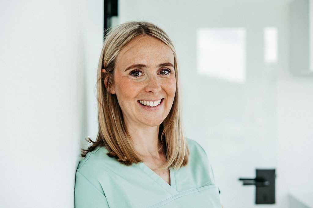 Dr. Alexandra Lampen, eine Frau mit schulterlangem, glattem, blondem Haar, steht in einem hellen, modern eingerichteten Praxisraum. Sie trägt grüne medizinische Kleidung und lehnt entspannt an einer weißen Wand. Sie lächelt freundlich in die Kamera. Im Hintergrund ist eine helle Tür mit schwarzem Griff zu sehen. Die Szene wirkt professionell und einladend – typisch für eine kieferorthopädische Praxis.