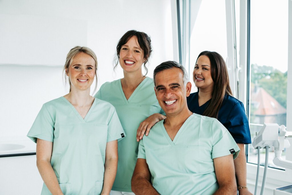 Vier Mitglieder des Praxisteams von U-Smile stehen und sitzen freundlich lächelnd in einem hellen Behandlungsraum mit großen Fenstern. Drei Frauen und ein Mann tragen moderne Kasacks in Mintgrün und Dunkelblau. Im Vordergrund sitzt ein Mitarbeiter, die anderen Teammitglieder stehen nah beieinander hinter ihm. Rechts im Bild ist ein Teil eines Intraoralscanners zu sehen, im Hintergrund fällt viel Tageslicht in die moderne kieferorthopädische Praxis.