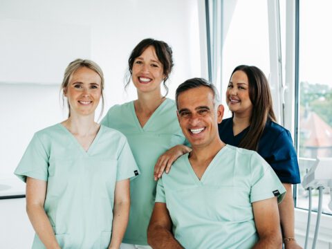 Vier Mitglieder des Praxisteams von U-Smile stehen und sitzen freundlich lächelnd in einem hellen Behandlungsraum mit großen Fenstern. Drei Frauen und ein Mann tragen moderne Kasacks in Mintgrün und Dunkelblau. Im Vordergrund sitzt ein Mitarbeiter, die anderen Teammitglieder stehen nah beieinander hinter ihm. Rechts im Bild ist ein Teil eines Intraoralscanners zu sehen, im Hintergrund fällt viel Tageslicht in die moderne kieferorthopädische Praxis.