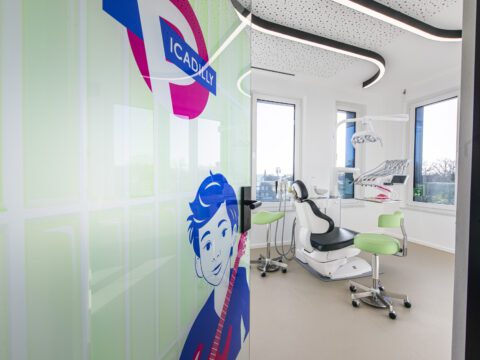 Modernes Behandlungszimmer der Kieferorthopädie U-Smile in Münster-Gremmendorf mit hellem, offenem Raumkonzept. Im Vordergrund ist eine Glastür mit einer Gestaltung im Londoner Tube-Stil zu sehen, inklusive rundem Symbol und der Aufschrift „Picadilly“ sowie einer comicartigen Figur in Blau und Pink. Dahinter befindet sich ein weißer kieferorthopädischer Behandlungsstuhl mit schwarzer Polsterung, grüne Hocker, eine Behandlungseinheit mit Instrumenten und große Fenster, die viel Tageslicht in den Raum lassen. Die geschwungene Deckenbeleuchtung unterstreicht das moderne, freundliche Praxisdesign.